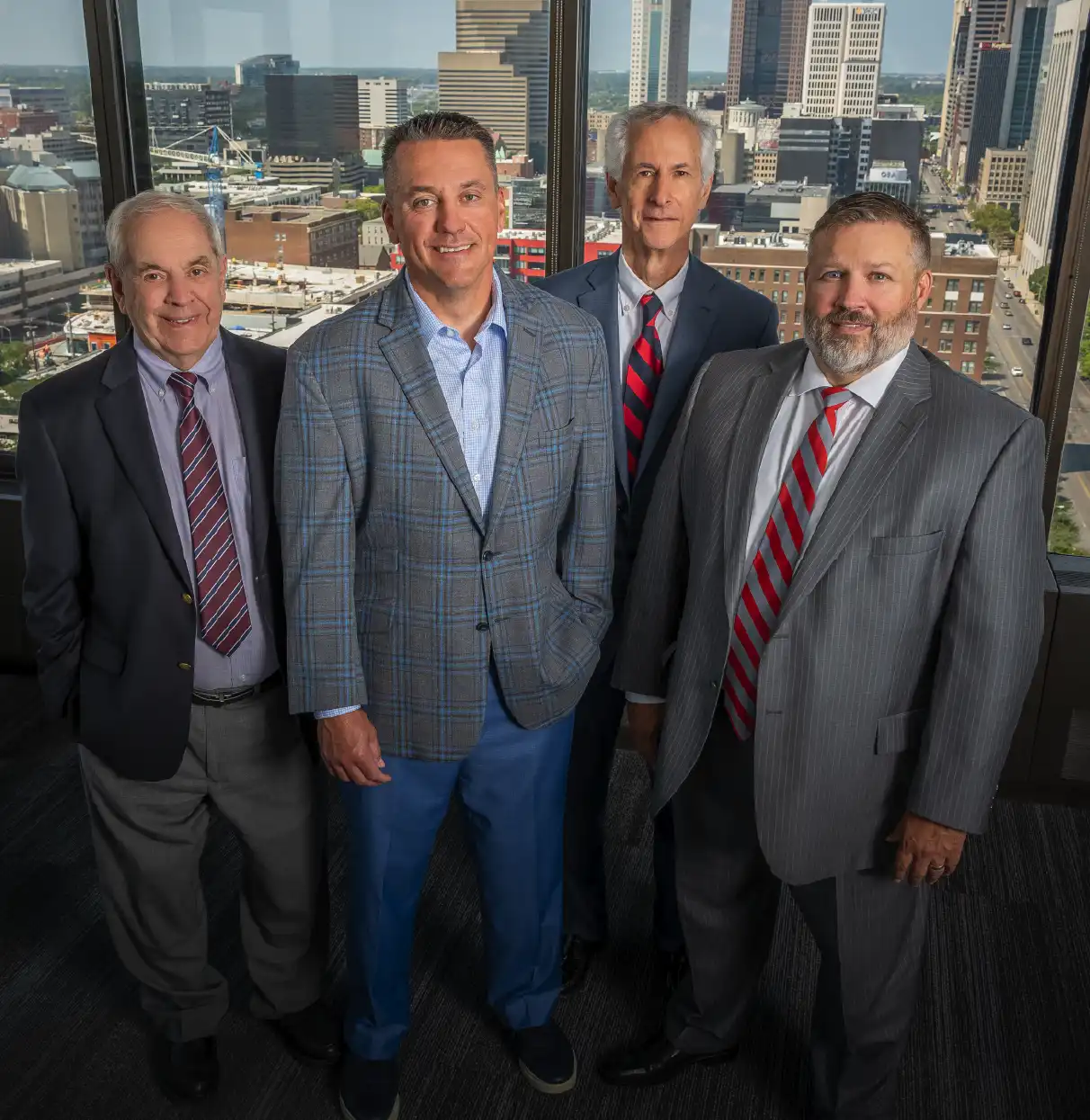 Four attorneys in business attire standing together in an office with a city skyline visible through the windows.